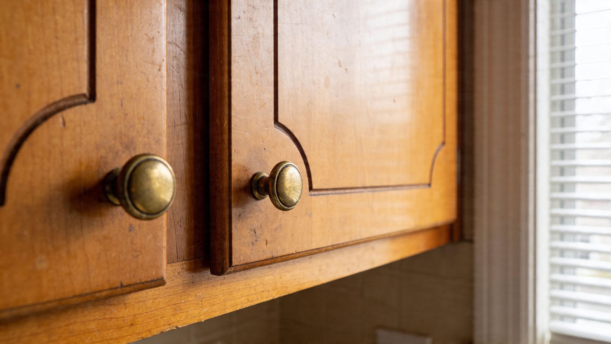 Close-up of classic wooden kitchen cabinets with brass knobs next to a window with blinds.