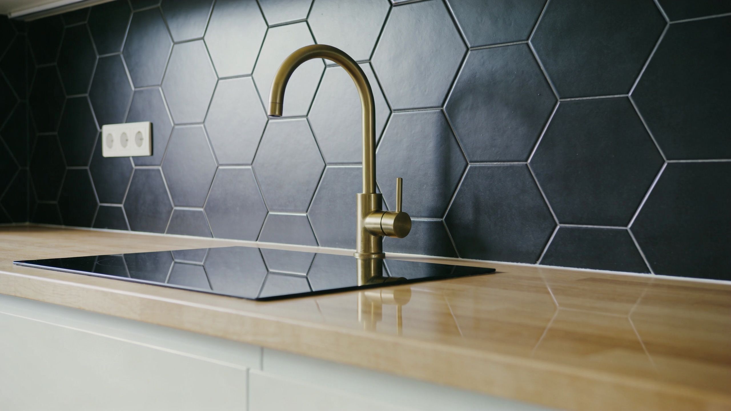 A modern kitchen counter with a sleek brass faucet set against a black hexagonal tile backsplash.